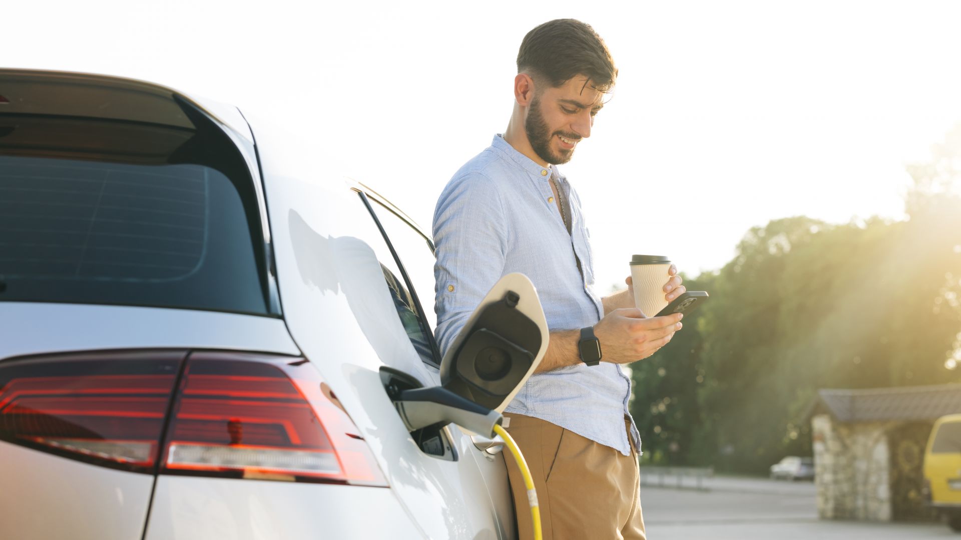 Man with smartphone and coffee stands with electric car at charging station